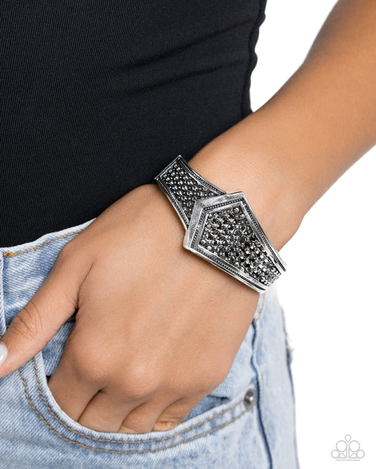 Order of the Arrow Silver Hinge Bracelet - Paparazzi Accessories A thick band of silver in the shape of an abstract arrow is framed by thick borders that line its edges. The interior surface is filled with tiny silver studs, and a sprinkle of hematite mixed in as the silver bar flares out in a dramatic finish. Features a hinged closure. Sold as one individual bracelet. P9ED-SVXX-137XX