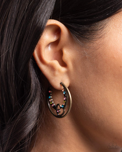 Sympathetic Sweetheart Brass Heart Hoop Earring - Paparazzi Accessories Glistening brass wire delicately bends into a heart frame from a smaller hoop inside a classic airy brass hoop, creating a flirtatious display. Dainty multicolored rhinestones are encrusted along the border of the daintier hoop and embellish a curve of the brass heart for additional romantic color and radiance. Earring attaches to a standard post fitting. Hoop measures approximately 1 1/4" in diameter. P5HO-BRXX-147XX