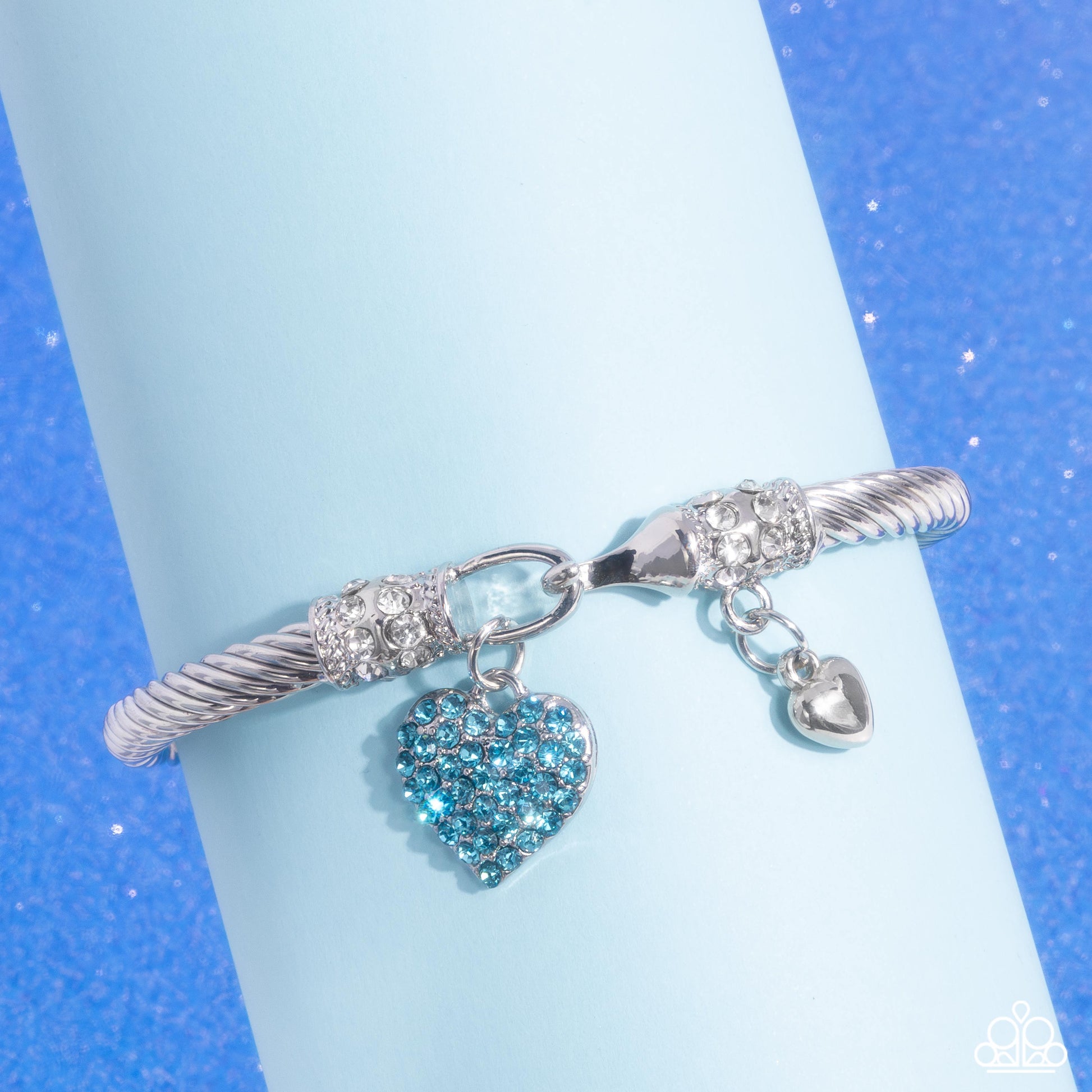 Heart Hour Blue Hook and Eye Bracelet - Paparazzi Accessories  Rippling with wavy textures, a glistening band of silver coils and twists along the wrist for a timeless display. An aquamarine rhinestone-encrusted silver heart swings from one end of the display while a dainty, solitaire silver heart swings from the other for a beautifully balanced look. Textured silver and rows of white rhinestones adorn the closures for additional refinement and glitz. Features a hook and eye closure.  Sold as one individual
