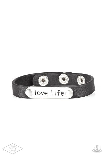 Love Life Black Wrap Bracelet - Paparazzi Accessories  A silver plate engraved with the inspirational phrase “love life” is studded in place along a skinny strip of black leather. Brushed in a shiny finish, the dainty band wraps around the wrist for a simple seasonal style. Features an adjustable snap closure.  Sold as one individual bracelet. This Fan Favorite is back in the spotlight at the request of our 2021 Life of the Party member with Black Diamond Access, Robert F.