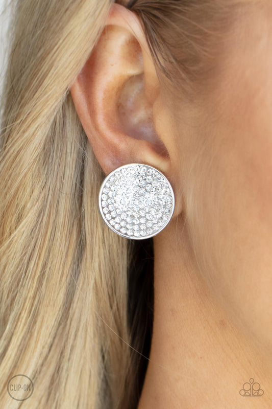Drama on Demand White Clip-On Earring - Paparazzi Accessories  Sparkling with brilliance, a concave shiny silver disc encompasses radiating rows of shimmering white rhinestones creating a striking impact. Earring attaches to a standard clip-on fitting.  Sold as one pair of clip-on earrings.