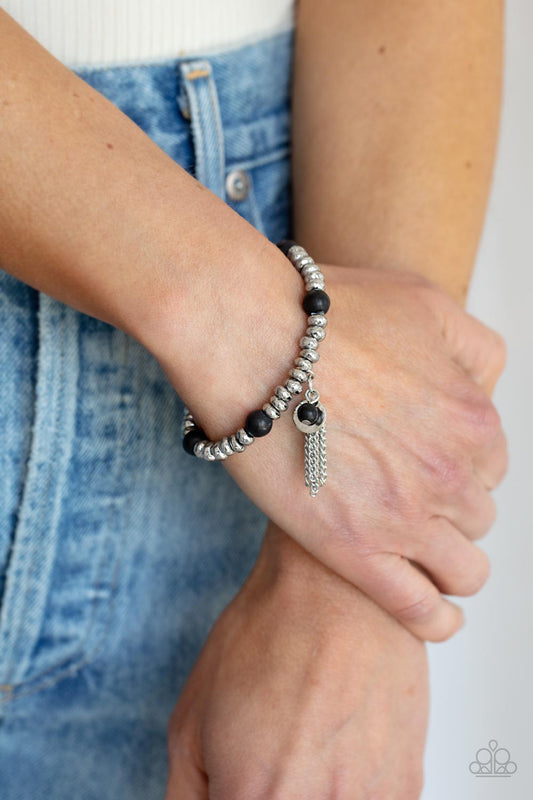 Whimsically Wanderlust Black Bracelet - Paparazzi Accessories  An earthy collection of hammered silver beads and round black stones are threaded along a stretchy band around the wrist. A dainty silver tassel, hammered silver disc, and a black stone swing from the seasonal display, adding playful movement to the whimsy look.  Sold as one individual bracelet.
