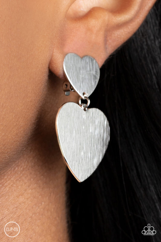 Cowgirl Crush Silver Clip-On Earring - Paparazzi Accessories  Featuring a shimmery scratched finish, an oversized silver heart charm swings from the bottom of a smaller silver heart charm, resulting in a flirtatious lure. Earring attaches to a standard clip-on fitting.  Sold as one pair of clip-on earrings.