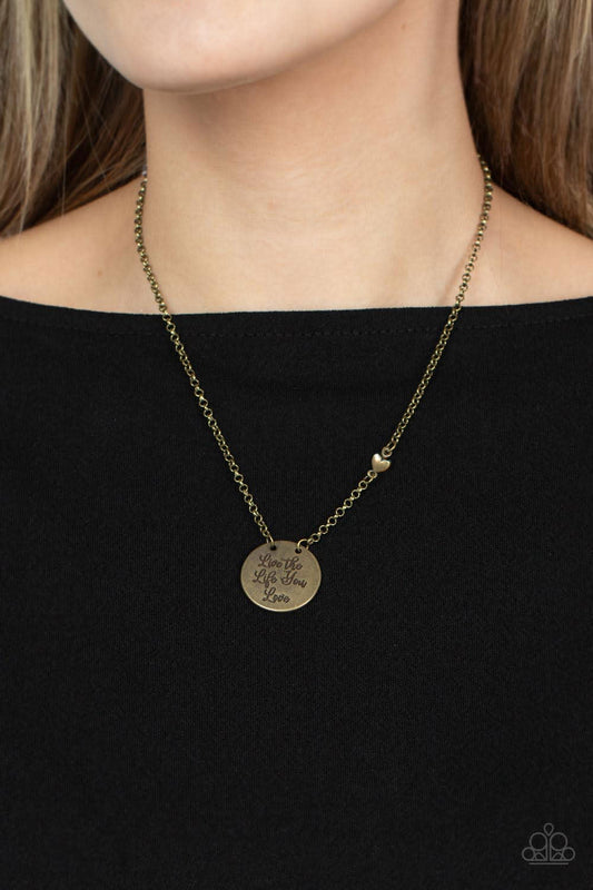 Live The Life You Love Brass Necklace - Paparazzi Accessories  A burnished brass disc is stamped in the inspiring phrase, "Live the Life You Love," at the bottom of a dainty brass chain, creating a motivational pendant. A dainty brass heart bead adorns one side of the chain for a light-hearted finish. Features an adjustable clasp closure.  Sold as one individual necklace. Includes one pair of matching earrings.