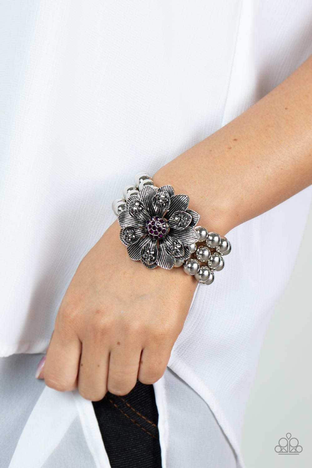 Botanical Bravado Purple Bracelet - Paparazzi Accessories  A daring oversized silver flower is composed of petals lined in antiqued silver and dotted with smoky hematite rhinestones. A sphere of dainty purple rhinestones creates the center of the flower as it sits atop a trio of shiny silver beaded stretchy bands around the wrist for a dramatically whimsical look.  Sold as one individual bracelet.