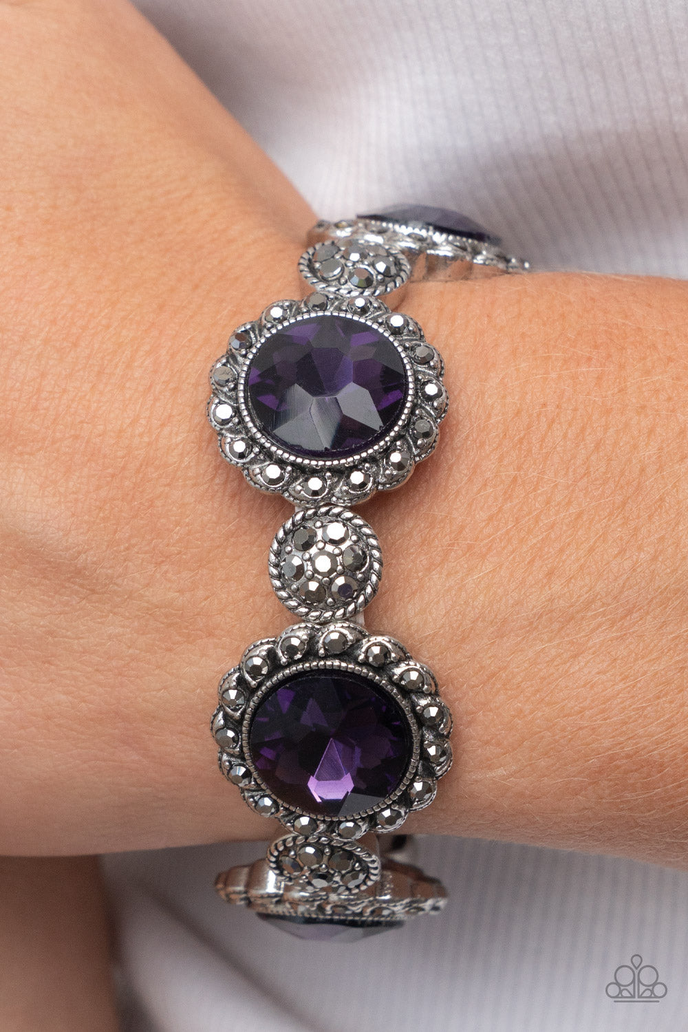 Palace Property Purple Bracelet - Paparazzi Accessories  An oversized purple rhinestone adorns the center of a hematite rhinestone petaled floral frame. Infused with hematite dotted silver accents, the glitzy floral frames sparkle along stretchy bands around the wrist for a glamorous finish.  Sold as one individual bracelet.
