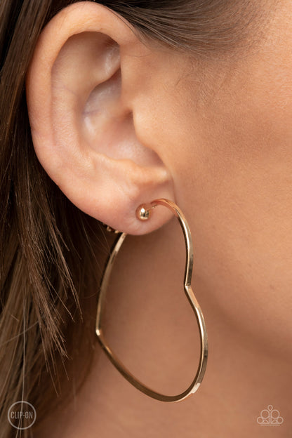 Harmonious Hearts Gold Clip-On Hoop Earring - Paparazzi Accessories  A glistening gold bar delicately curves into an oversized heart frame, resulting in a heart-stopping shimmer. Earring attaches to a standard clip-on fitting.  Sold as one pair of clip-on earrings.