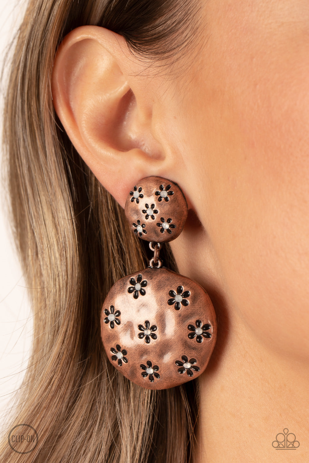 Industrial Fairytale Copper Clip-On Earring - Paparazzi Accessories  Dotted with dainty opal rhinestone centers, an antiqued floral pattern is stamped across the fronts of two hammered copper discs that delicately connect into a rustic lure. Earring attaches to a standard clip-on fitting.  Sold as one pair of clip-on earrings.