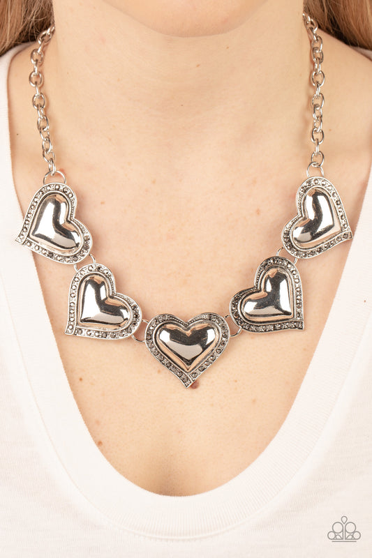 Kindred Hearts Silver Necklace - Paparazzi Accessories  Bordered in glitzy hematite rhinestones, an oversized collection of beveled silver heart frames delicately links below the collar for a dramatically romantic look. Features an adjustable clasp closure.  Sold as one individual necklace. Includes one pair of matching earrings.