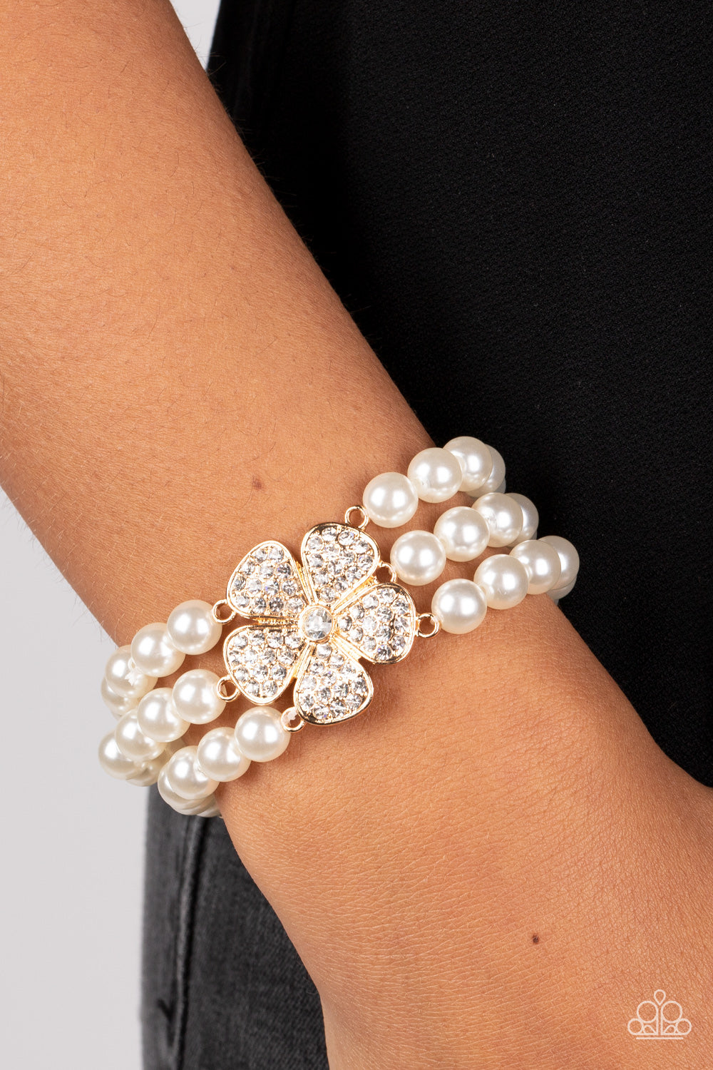 Park Avenue Orchard Gold Pearl Bracelet - Paparazzi Accessories  Separated by dainty gold plates, classic white pearls are threaded along three stretchy bands around the wrist. Encrusted in glassy white rhinestones, a glitzy gold flower blooms at the center of the wrist for a fierce floral statement.  Sold as one individual bracelet.