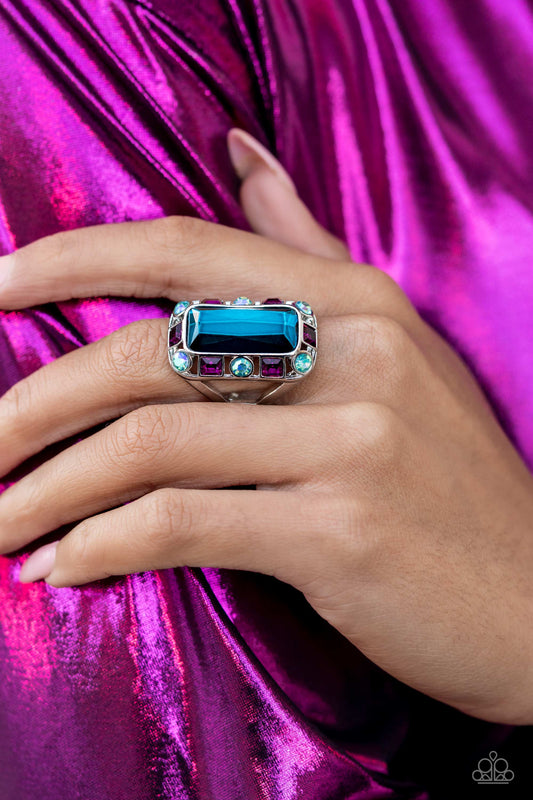 Radiant Rhinestones Blue Ring - Paparazzi Accessories  Pressed into a curved, rectangular silver frame, an oversized emerald-cut blue gem shines atop the finger. Dainty square and round gems in shades of fuchsia and iridescent blue airily encircle the oversized gem for some additional shimmer resulting in a radiant centerpiece. Features a stretchy band for a flexible fit. Due to its prismatic palette, color may vary.  Sold as one individual ring.  ﻿﻿December 2022 Life of the Party!