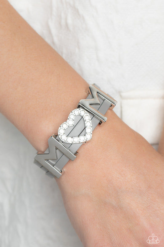 Heart of Mom Silver Magnetic Bracelet - Paparazzi Accessories  A row of glassy white rhinestones are encased into a sparkly heart frame that is threaded along layers of gray leather strands around the wrist. Framing both sides of the heart frame, two high-sheen silver letter "M"s spell out the word "mom" atop the wrist for a sentimental finish. Features a magnetic closure.  Sold as one individual bracelet.  P9WD-SVXX-218XX