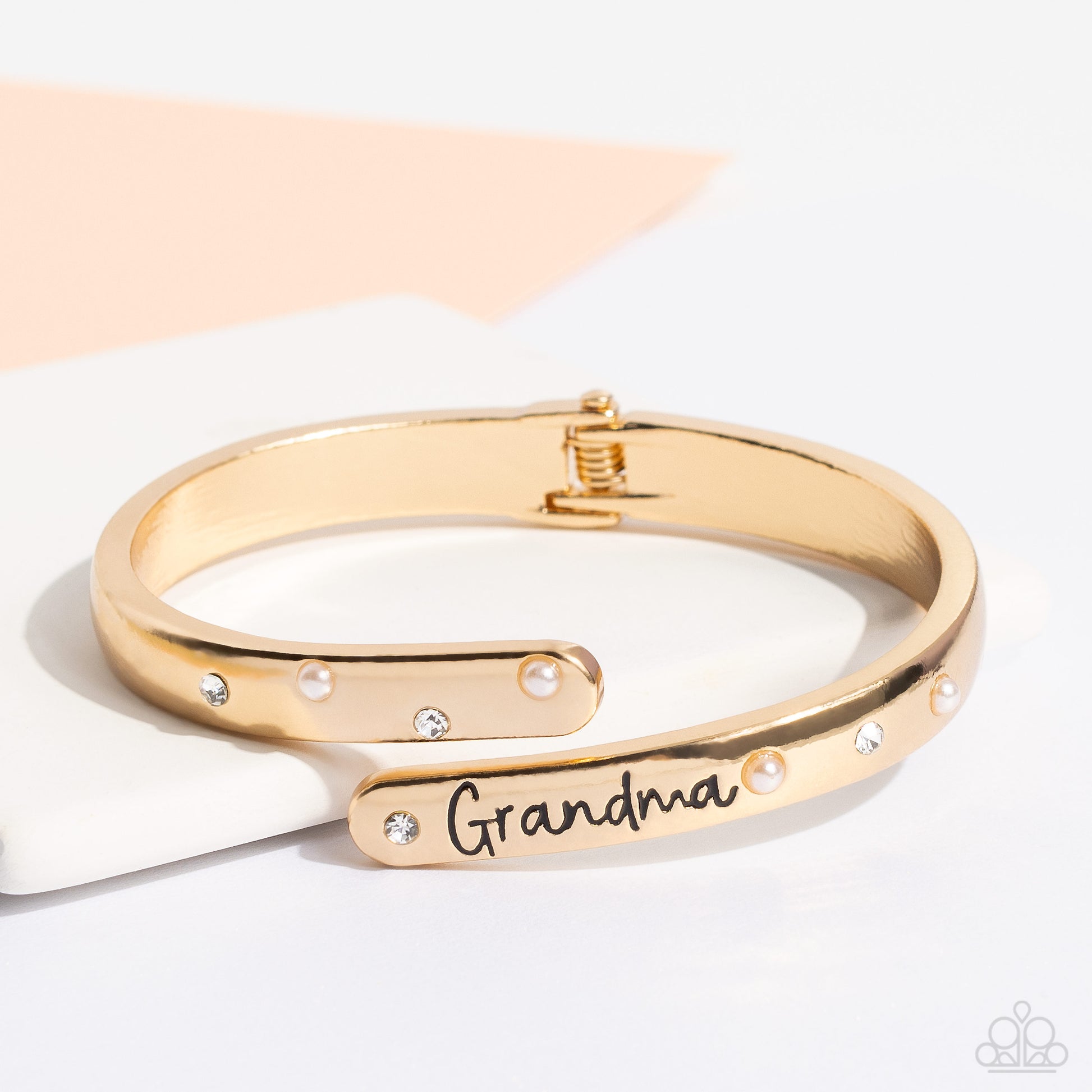 Gorgeous Grandma Gold Hinge Bracelet - Paparazzi Accessories  High-sheen gold curves into a hinged closure around the wrist. Featured on the upper curve, dainty pearls and white rhinestones alternate in a zig-zag pattern for a refined shimmer. The lower curve of gold features the stamped word "Grandma" in a curly font with rhinestones and pearls bordering the word for additional elegance. Features a hinged closure.  Sold as one individual bracelet.  P9WD-GDXX-191XX