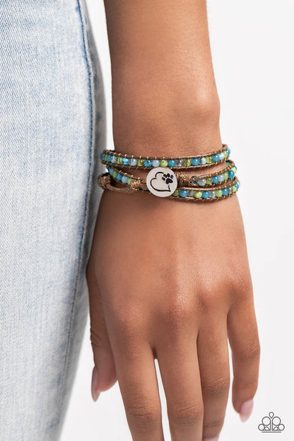PAW-sitive Thinking Blue Wrap Bracelet - Paparazzi Accessories  Infused with an antiqued heart and pawprint embossed button, a row of blue and green multicolored cloudy and milky beads are knotted in place along two brown leather cords, creating multiple earthy layers around the wrist with its extended length. Features an adjustable button loop closure.  Sold as one individual bracelet.  P9UR-BLXX-254XX
